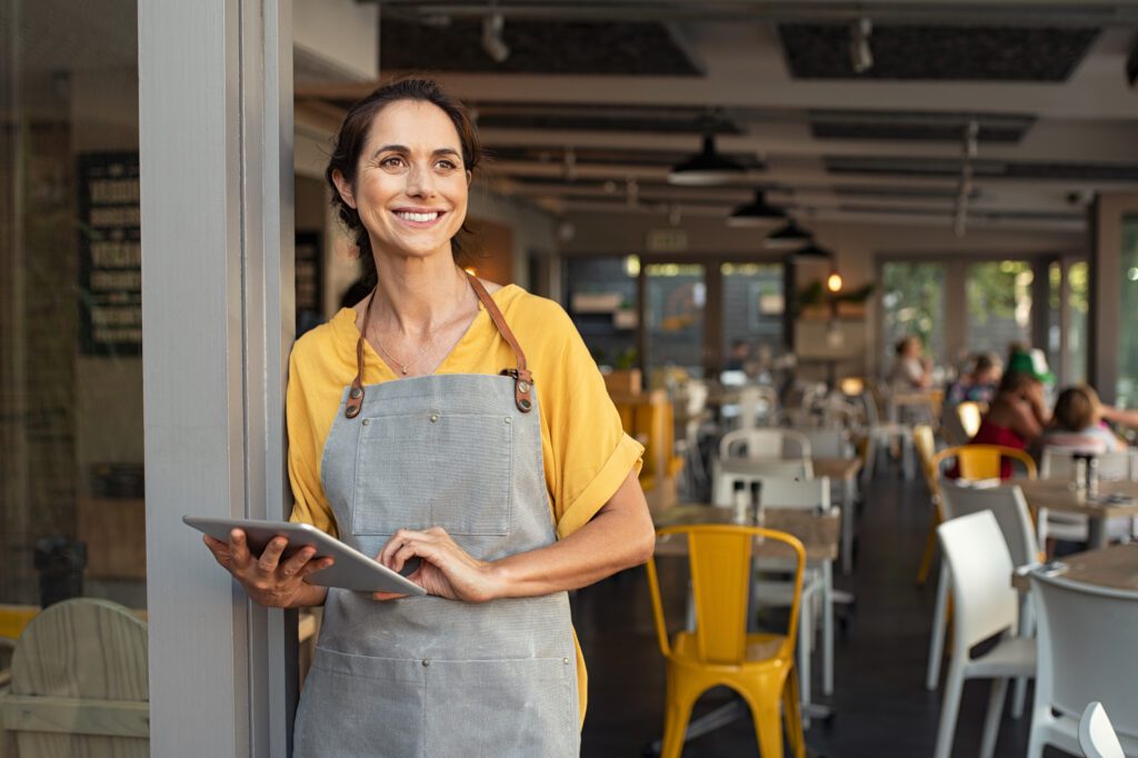 A woman in an apron holding a tablet and smiling at the entrance to her business.
