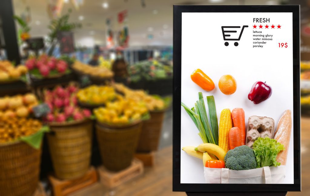 A digital sign promoting fresh produce in front of fruits and vegetables in a grocery store.