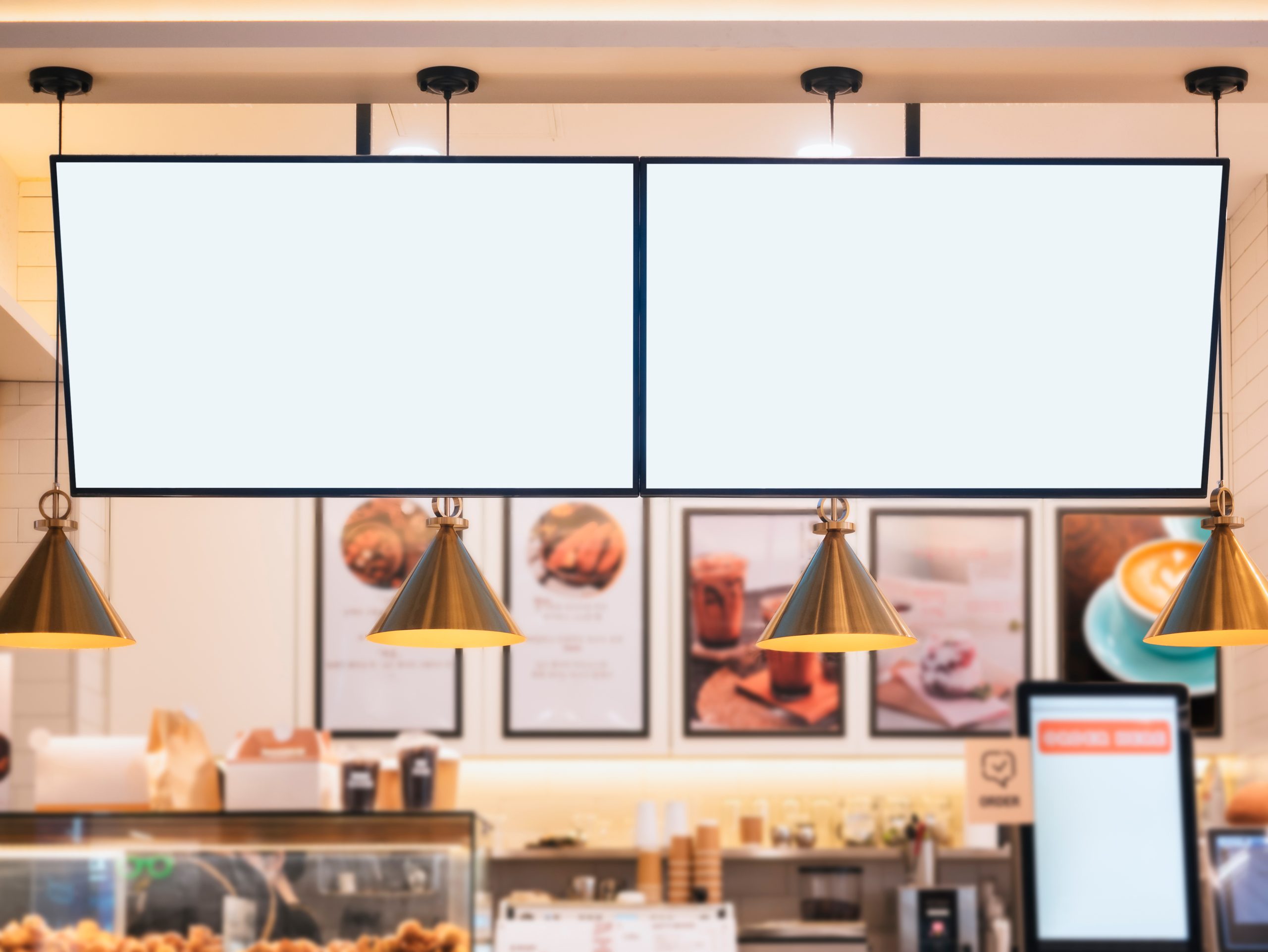 Two large digital screens are hung over a café counter, with analog signage behind the counter.
