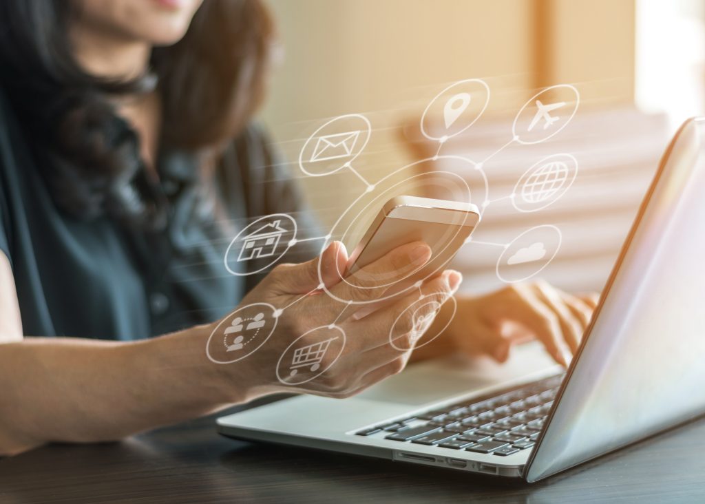 A person at their laptop with a phone in hand, with multiple icons displayed over their phone.