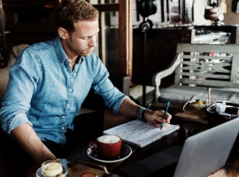 Young entrepreneur working on business project in city cafe, writing down ideas and using laptop
