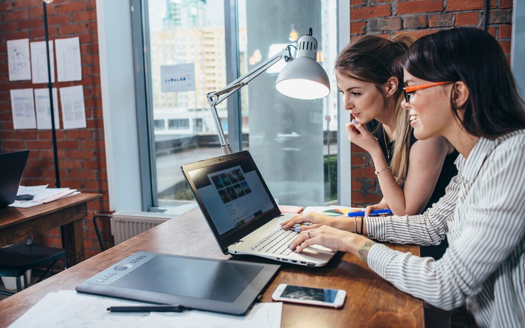 A pair of women at a small wooden table look over a laptop and tablet.