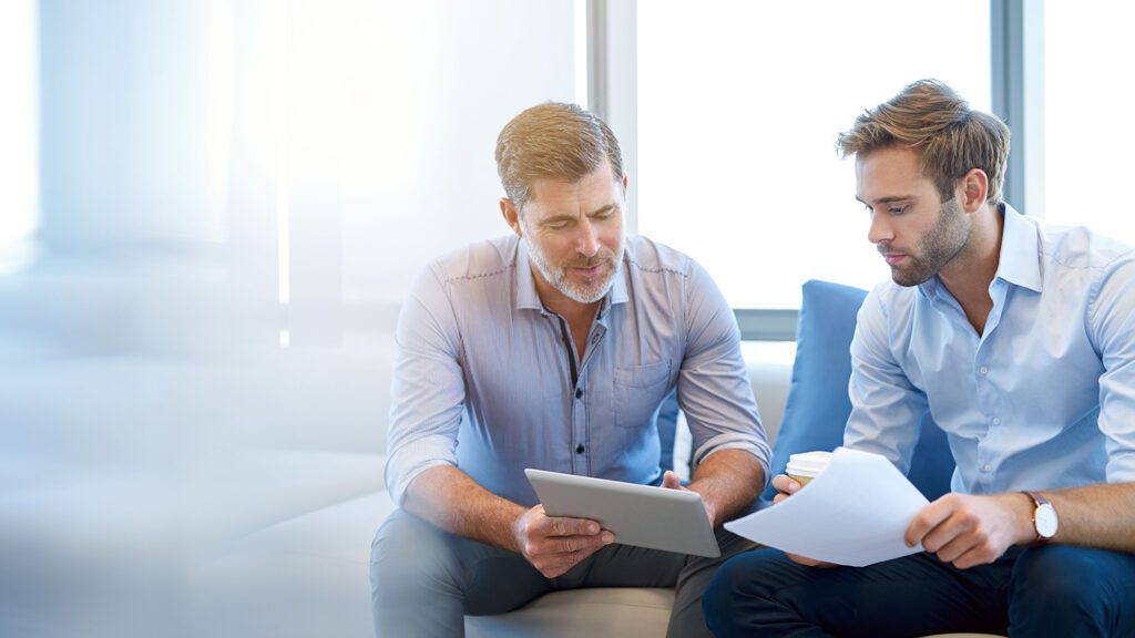 An older businessman speaks to his younger colleague while pointing to a tablet.